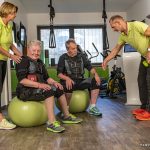 Zwei Senioren sitzen auf grünen Gymnastikbällen und trainieren mit Elektrostimulationsanzügen in einem Fitnessstudio.