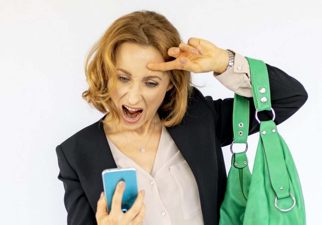 Eine Frau in eleganter Bürokleidung schaut überrascht oder erfreut auf ihr Smartphone, während sie eine grüne Handtasche über der Schulter trägt.